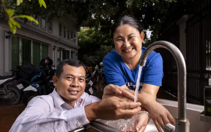 A man and a woman look at the camera, they are washing their hands together at a sink