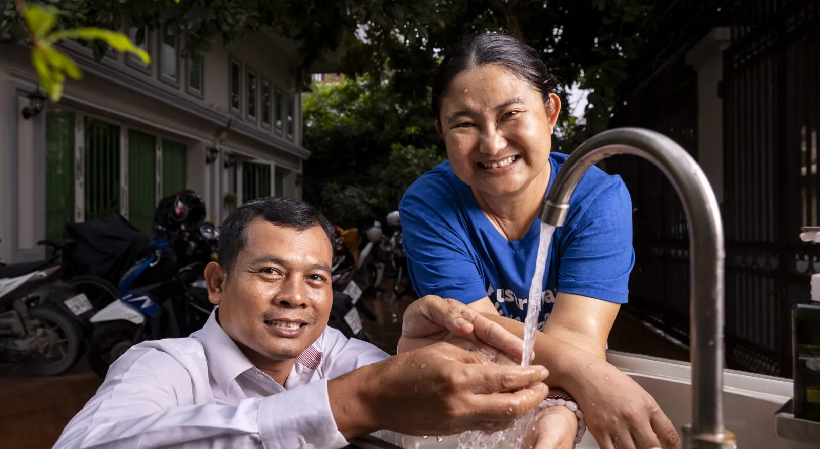 A man and a woman look at the camera, they are washing their hands together at a sink