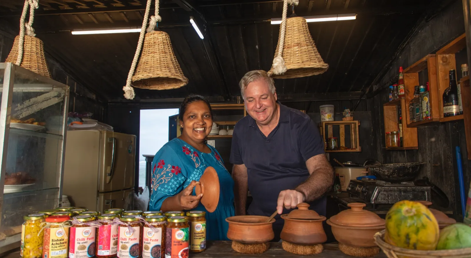 A man and a woman stand behind a table which has food and drinks on it. Both are smiling.