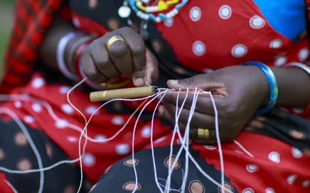 A close up of a lady's hands as she works on beading. She is wearing a bright red and white outfit and a colourful necklace