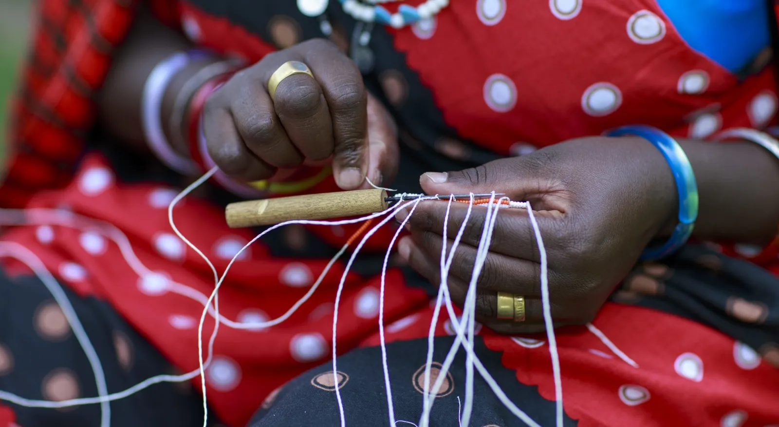 A close up of a lady's hands as she works on beading. She is wearing a bright red and white outfit and a colourful necklace