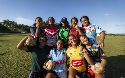 A group of seven teenagers and an older lady smile at the camera and hug each other. They are all wearing rugby uniforms of different colours and some hold rugby balls.