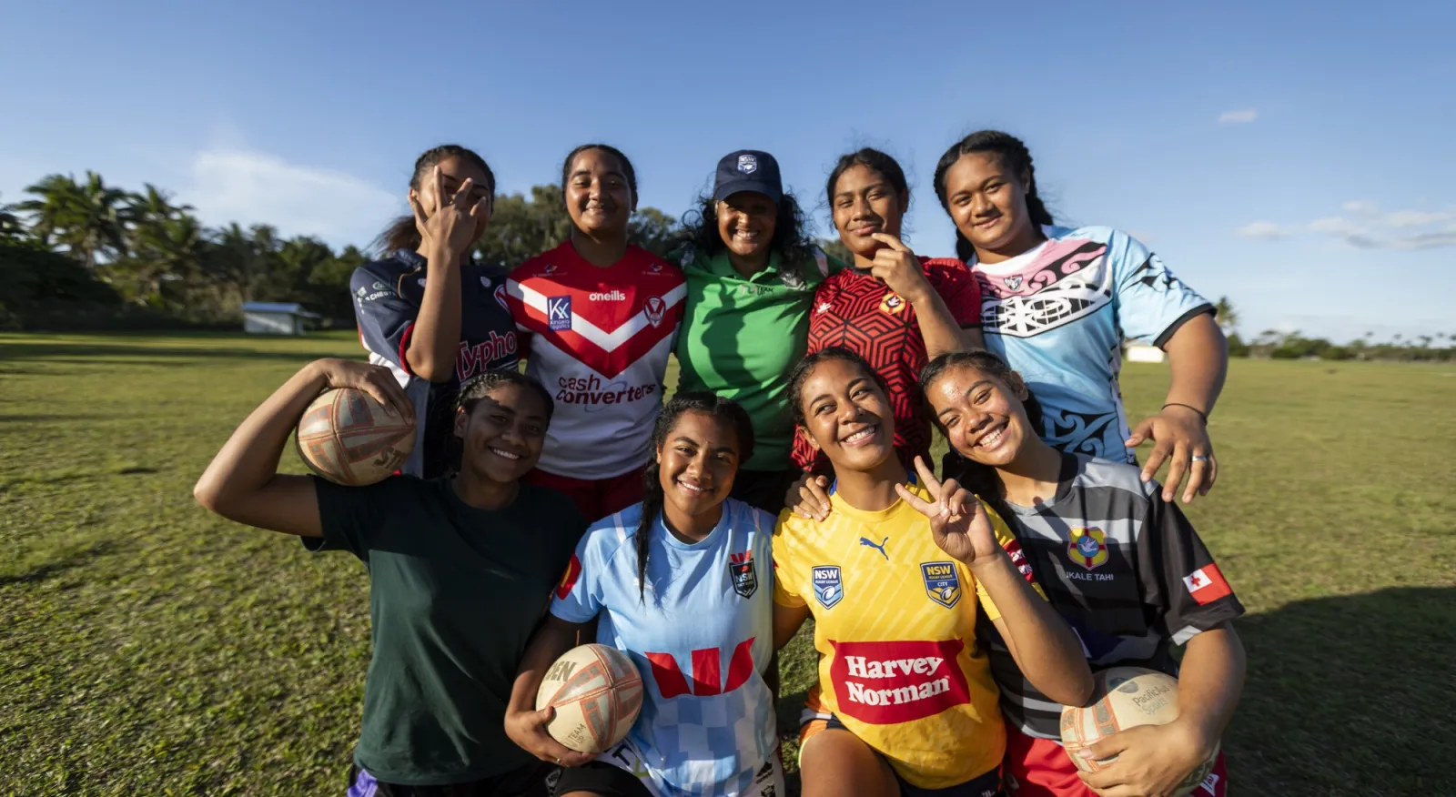 A group of seven teenagers and an older lady smile at the camera and hug each other. They are all wearing rugby uniforms of different colours and some hold rugby balls.