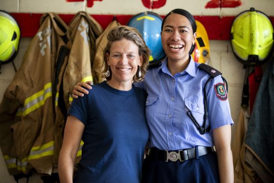 Two women look into camera and smile, one wears a uniform. Behind them are firemen's/firewomen's uniforms hanging up.