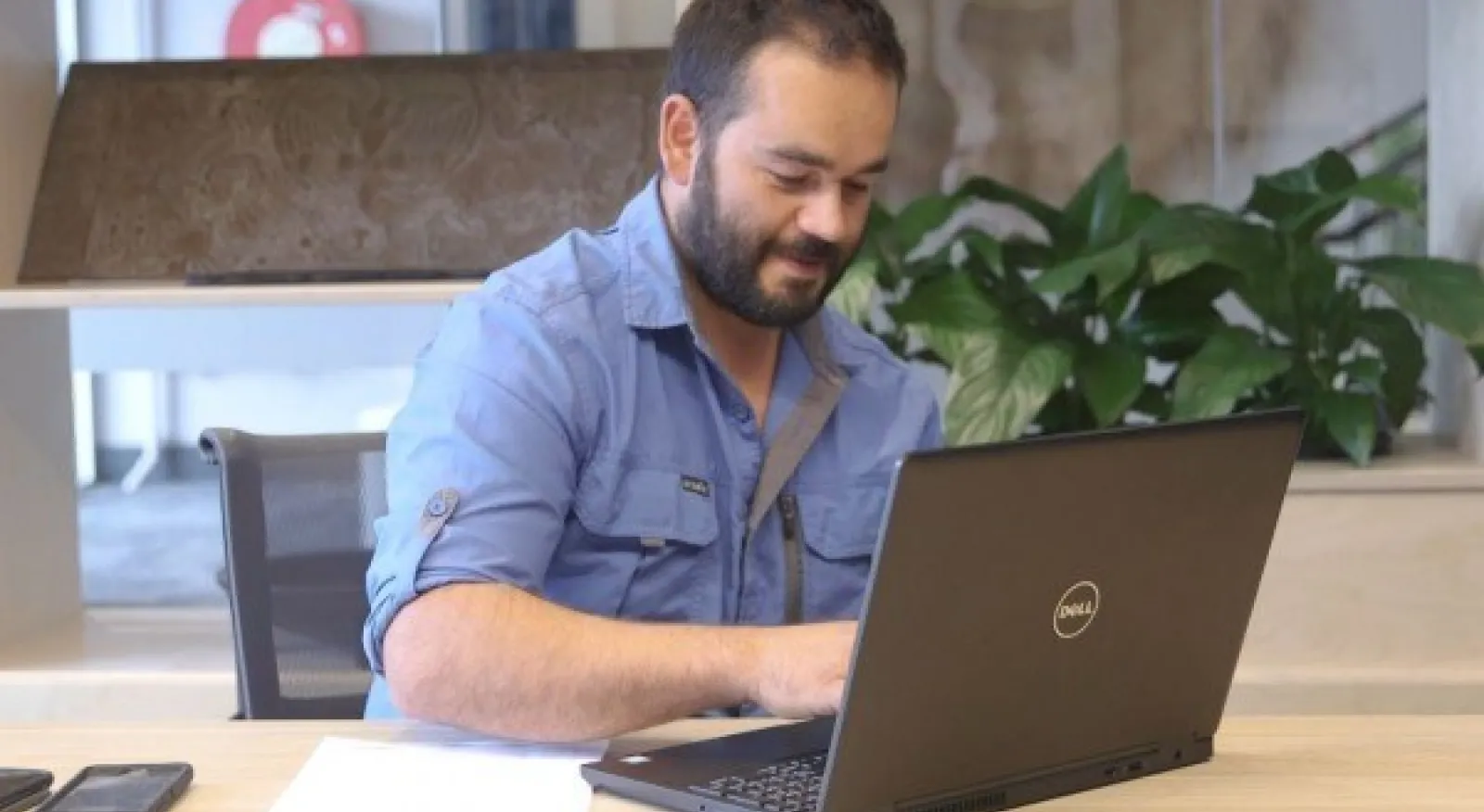 Clinton, a young man in a blue shirt, sits in front of a laptop in an office. He is typing and looking at the laptop screen.