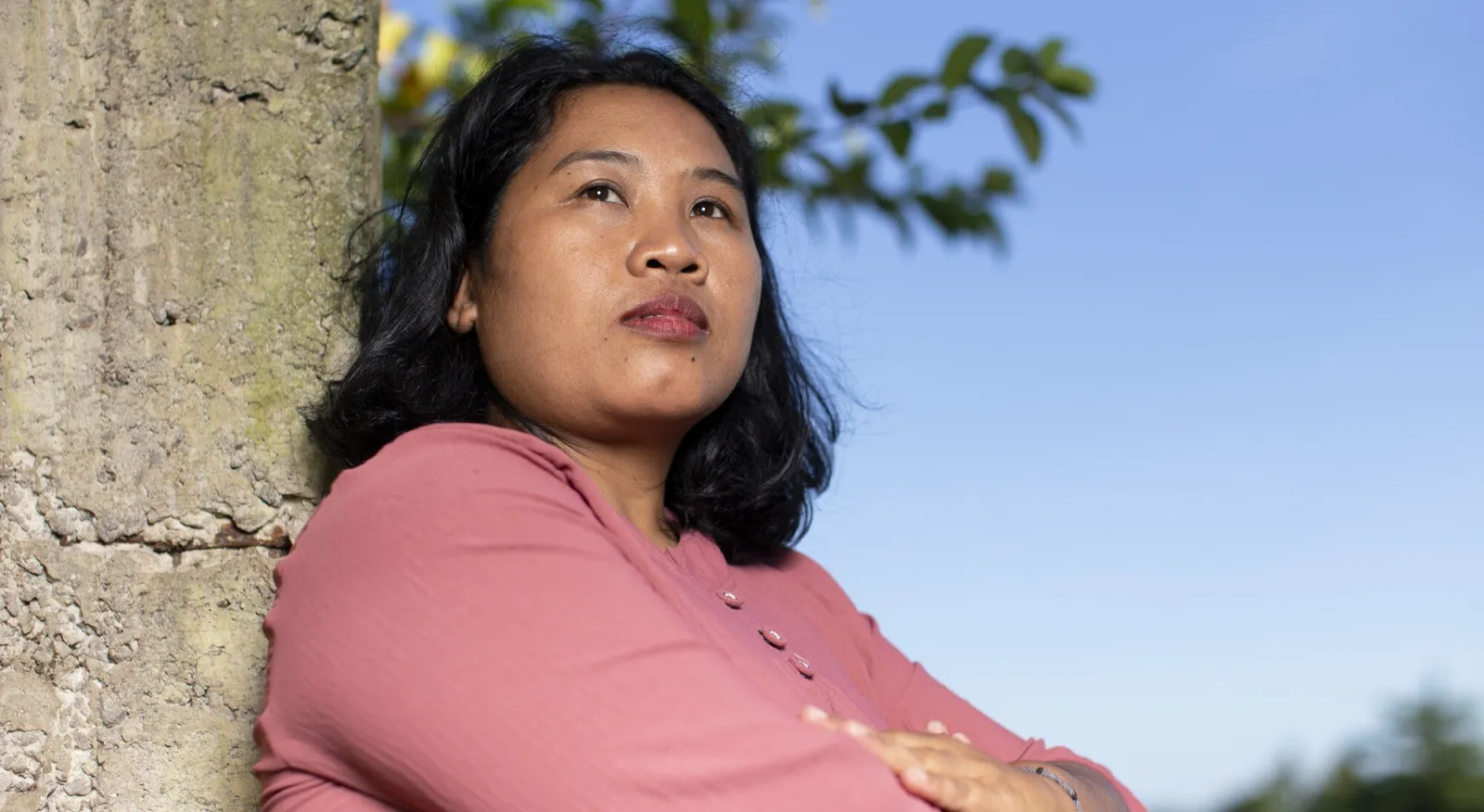 A woman is folding her arms and looking into the distance with a serious expression. She is leaning against a tree with a blue sky backdrop. She has a pink shirt and shoulder length black hair.