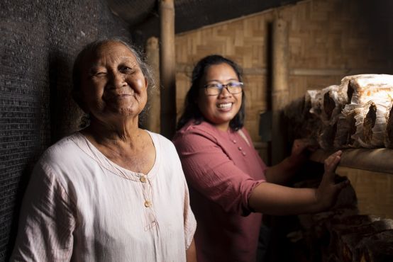 Tow women smiling at the camera. The woman in the foreground is elderly and wears a white top, while the woman behind her has a pink top. They are in a storeroom.
