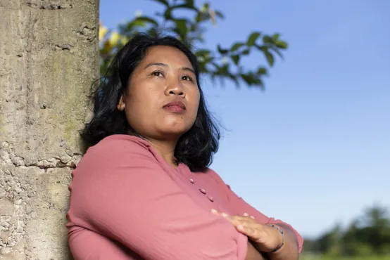 A woman is folding her arms and looking into the distance with a serious expression. She is leaning against a tree with a blue sky backdrop. She has a pink shirt and shoulder length black hair.