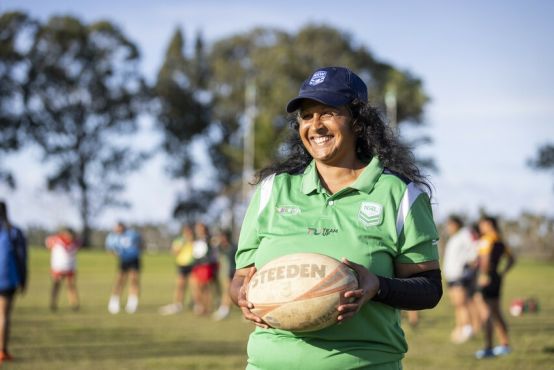 A woman holding a rugby ball smiling on a rugby field with female players in the distance.